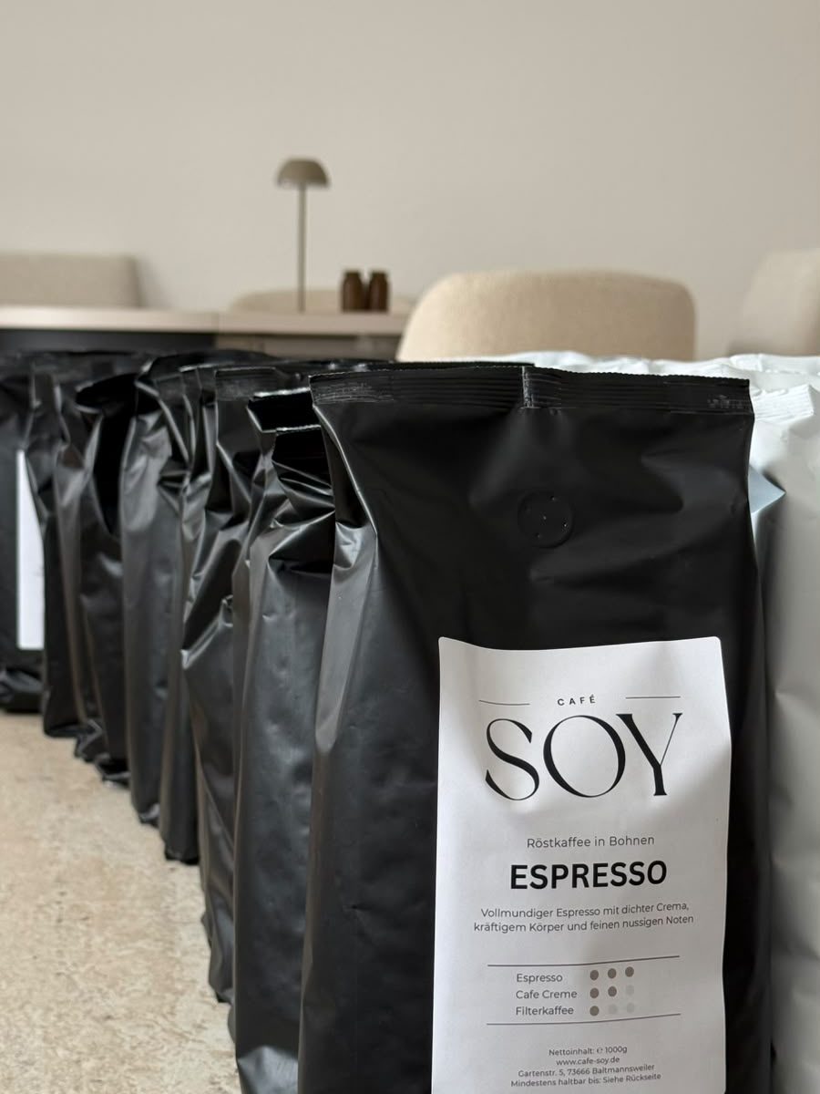 Eine Reihe schwarzer Espresso-Tüten von Café SOY auf einem Tisch, dahinter Stühle und eine Tischlampe